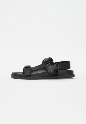 Black leather sandals with adjustable straps, a flat sole, and cut-out design details for breathability. Features stitching accents.