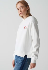 Felpa bianca con cappuccio e tasca frontale, dettaglio del logo in rosso e grigio, vestibilità comoda, polsini e orlo a coste, abbinata a jeans in denim blu.