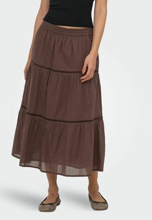 Femme portant une jupe midi marron à volants avec une ceinture élastique et un haut noir, debout, les mains détendues le long du corps, portant des ballerines marron.