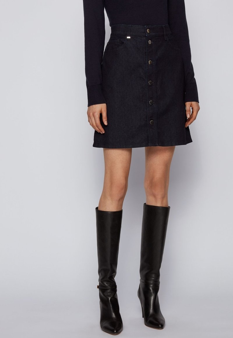 Femme portant une jupe en jean noire à boutons devant, un haut noir à manches longues, et des bottes en cuir noires montantes jusqu'aux genoux, sur un fond uni.
