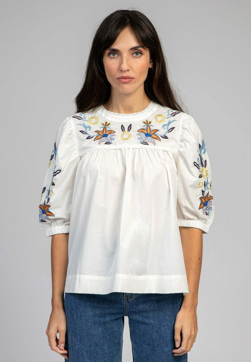 Blouse en coton blanc avec des motifs floraux brodés en bleu, orange et jaune. Dotée de manches bouffantes et d'un col rond.