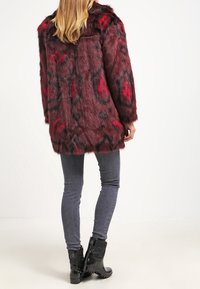 Abrigo de piel sintética en rojo oscuro con patrones en negro y carmesí, con cuello redondeado y mangas largas, combinado con jeans ajustados oscuros y botas negras.