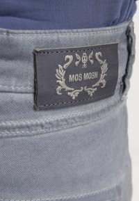 Šedá tkanina s texturovaným povrchem, na kterém je tmavě modrá etiketa s nápisem "MOS MOSH" a dekorativními akcenty.