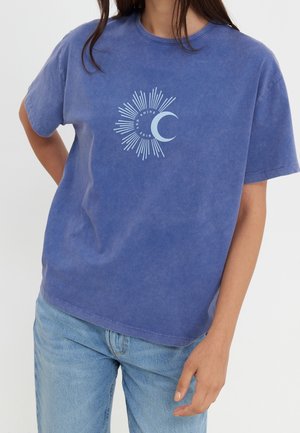 Kobieta ubrana w wyblakły niebieski t-shirt z białą grafiką promieni słonecznych i półksiężyca oraz napisem „RISE AND SHINE”, w połączeniu z jasnoniebieskimi jeansami.