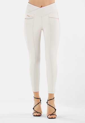 Nainen, jolla on korkeavyötäröiset, luonnonvalkoiset istuvat housut, joissa on etutaskut vetoketjuilla, ja mustat, remmillä varustetut korkokengät.