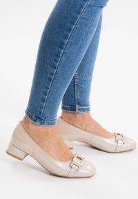 Chaussures à talons bas en cuir verni beige avec un détail de boucle carrée au niveau des orteils, portées avec un jean skinny bleu, mettant en valeur une texture lisse.
