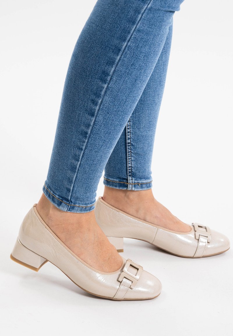 Chaussures à talons bas en cuir verni beige avec un détail de boucle carrée au niveau des orteils, portées avec un jean skinny bleu, mettant en valeur une texture lisse.
