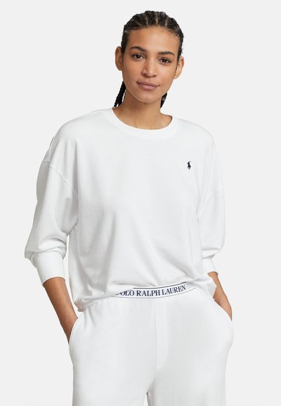 Νεαρό άτομο με κοτσίδες, φορώντας λευκό πουκάμισο μακρυμάνικο και λευκό παντελόνι με τη ζώνη μέσης με το λογότυπο Polo Ralph Lauren, με τα χέρια στις τσέπες.