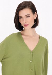 Zöld V nyakú kardigán, puha kötött anyagból készült, öt azonos gombbal és hosszú ujjakkal. A modellen karika fülbevalók találhatók.
