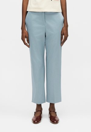 Osoba stojaca proti jednému bielemu pozadiu, oblečená v svetlomodrých nohaviciach širokého strihu, v krémovom topu a tmavočervených topánkach Mary Jane.