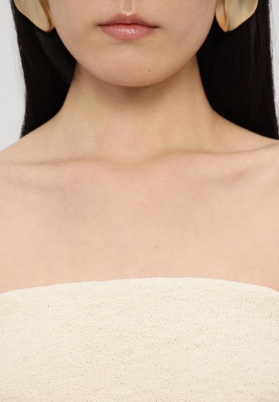 Beige trägerloses Oberteil, strukturierter Stoff mit lockerem Gewebe. Glatte Haut und große, cremefarbene Statement-Ohrringe, minimalistisches Design.