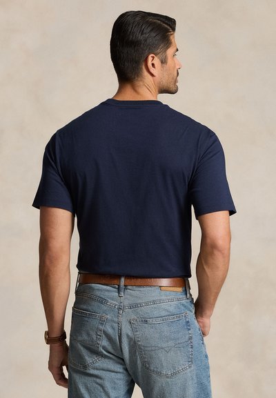 Mann iført en marineblå t-skjorte med korte ermer, stukket inn i lyseblå jeans med et brunt lærbelte, med ryggen mot kameraet.