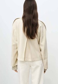 Beige oversized jacka med en strukturerad design, som har en plisserad rygg och avslappnade ärmar. I kombination med off-white byxor.