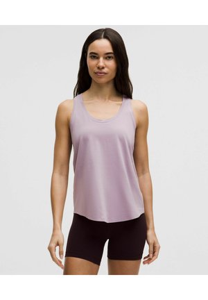 Kvinde iført en lys lilla ærmeløs tanktop og sorte tætsiddende shorts, stående mod en ensfarvet lys grå baggrund.