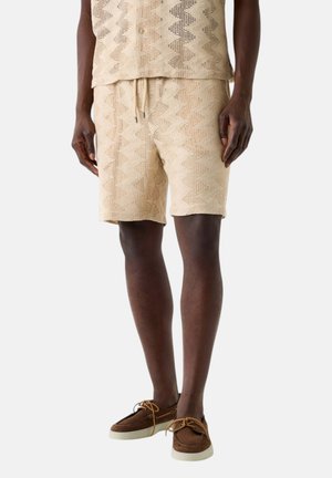 Benen van een persoon die beige korte broek met patroon en bijpassend shirt draagt, gecombineerd met bruine leren bootschoenen met witte zolen en veters.