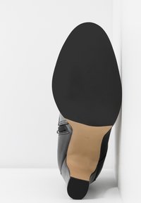 Bottine en cuir noir inclinée vers le haut montrant une semelle noire lisse et un marquage de taille 37 sur une semelle intermédiaire beige, sur fond blanc.