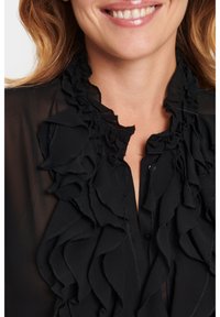 Blusa nera con colletto arricciato e patta frontale, realizzata in tessuto trasparente con una texture liscia e una leggera lucentezza.