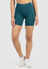 Model in teal hoge taille sport shorts en witte sneakers, staand voor een effen lichte achtergrond, die de pasvorm van de outfit toont.