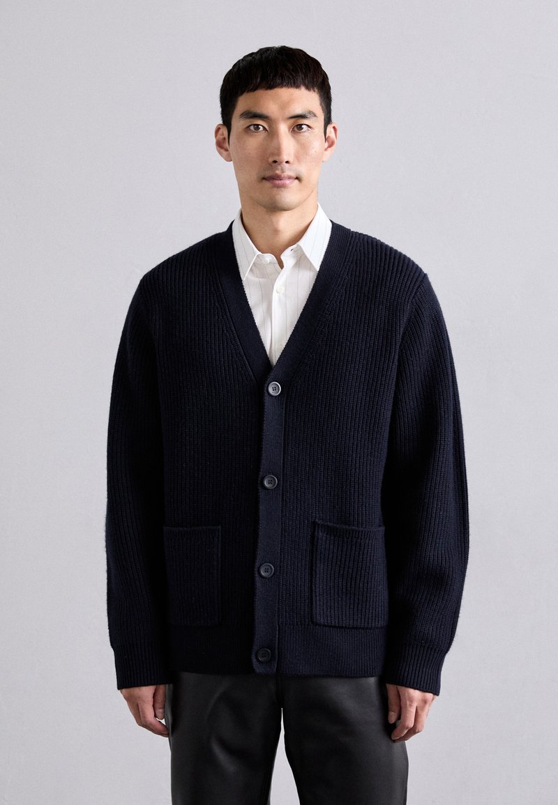 Tumesinine tekstuuriga kardigan V-kaelusega, viie nupuga ja kahe esitäpiga. Kangas näeb välja paks ja soe ning sellel on soonikdisain.