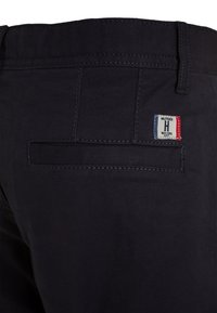 Pantaloni di un blu scuro con una texture liscia, con una tasca posteriore di forma rettangolare e un'etichetta con logo in fili rossi, bianchi e blu.