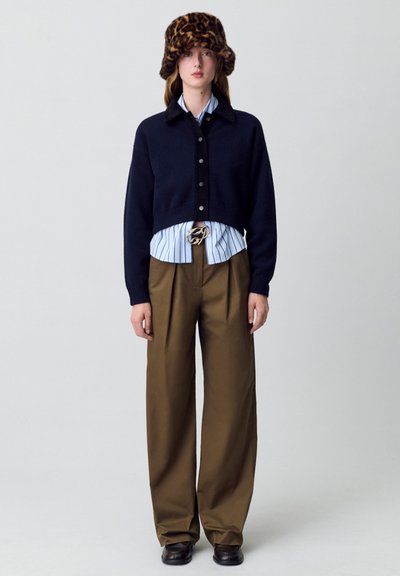 Suéter navy recortado con botones, camisa a rayas azul y blanca debajo, pantalones anchos de talle alto con pliegues marrones y un sombrero de estampado de leopardo marrón.