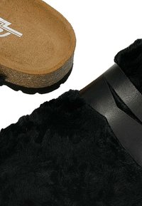 Černé pantofle na klínu s texturovaným povrchem připomínajícím kožešinu a hladkým černým koženým páskem. Podešev je z korku s vzorovaným protiskluzovým vzorkem.