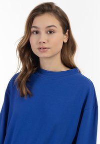 Kék, túlméretezett kapucnis pulóver széles nyakkivágással és eső vállakkal, puha, strukturált anyagból készült, hosszú ujjakkal és minimális varrással.