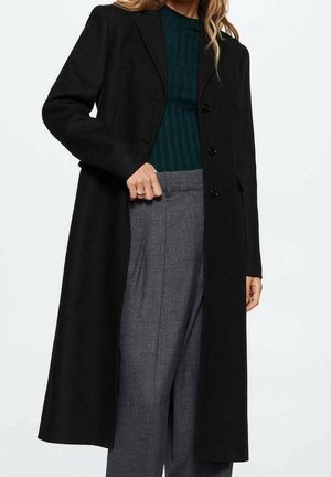 Vrouw met een lange zwarte jas met knopen over een donkergroene geribde trui en hooggetailleerde grijze geplooide broek, hand rustend bij de taille.