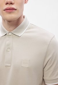 Μπεζ πόλο πουκάμισο από υφασμένο βαμβάκι με λευκό γιακά και διακόσμηση. Διαθέτει μικρό τετράγωνο λογότυπο με τη λέξη "BOSS" στο αριστερό μέρος.