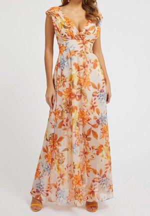 Vrouw in een mouwloze, V-hals, lange bloemenjurk met oranje, gele en blauwe bloemen op een lichte achtergrond, gecombineerd met oranje sandalen.