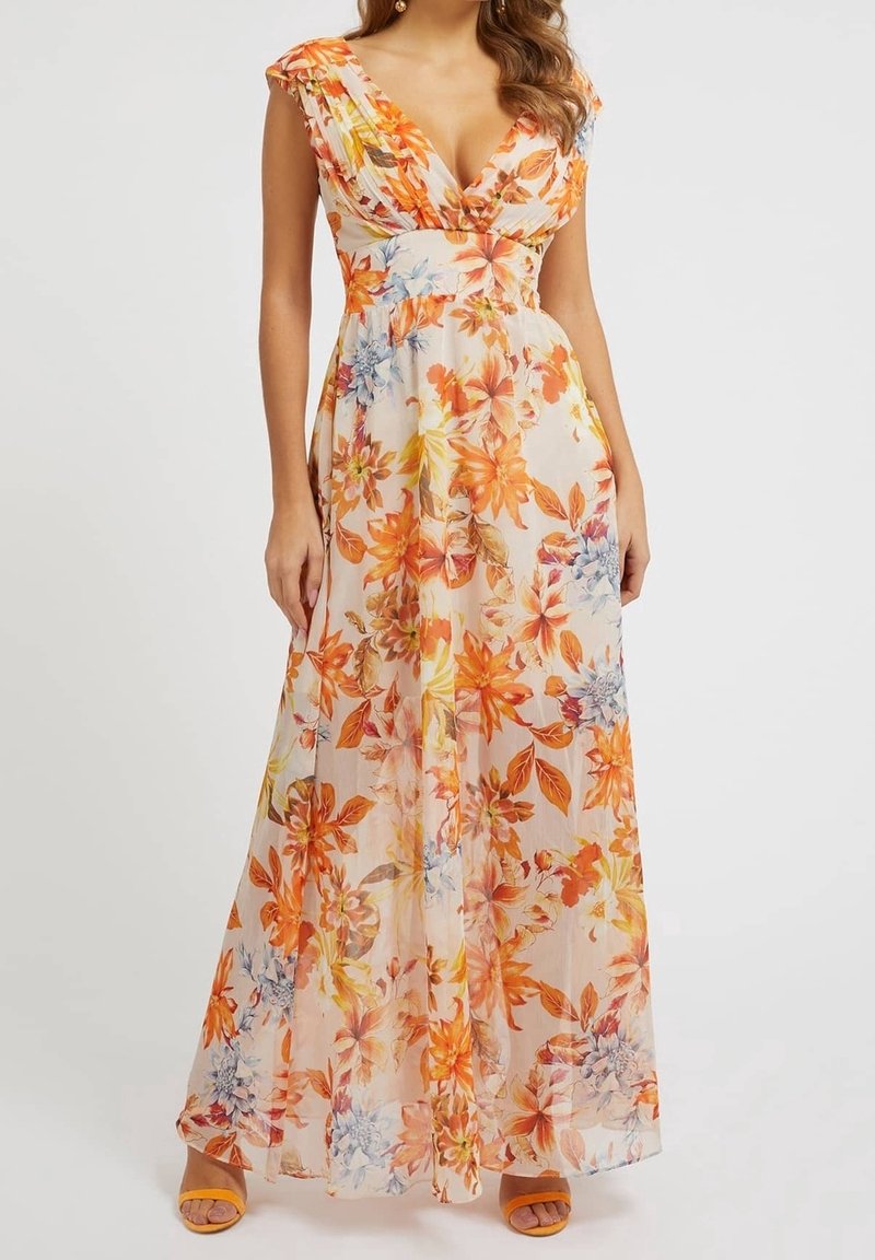 Femme portant une robe longue sans manches à col en V, à fleurs orange, jaune et bleues sur un fond clair, assortie à des sandales orange.