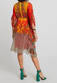 Abito con stampa floreale caratterizzato da un vivace top arancione e giallo, strati grigi trasparenti con pois e sandali con tacco verde smeraldo.