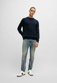 Marineblauwe gebreide trui met een ronde hals en geribde manchetten, gecombineerd met lichtblauwe faded jeans en witte sneakers met zwarte accenten.