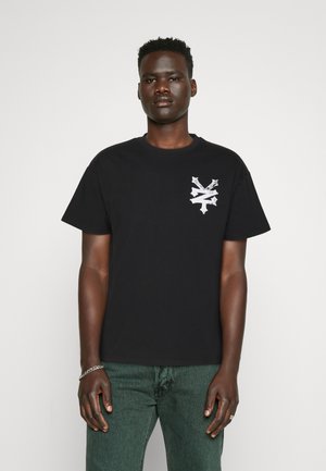 Sort bomulds T-shirt med et hvidt grafisk design på venstre bryst, korte ærmer og rund halsudskæring. Bæres med grønne bukser.