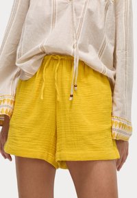 Pantalones cortos amarillos texturizados con cinturilla ajustable y dos cuentas de acento; combinados con una camiseta de rayas de manga larga que presenta detalles bordados.