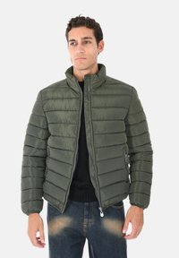 Giacca a vento verde con colletto alto, chiusura con zip e design trapuntato. Abbinata a jeans scuri. Tessuto liscio, isolamento caldo.
