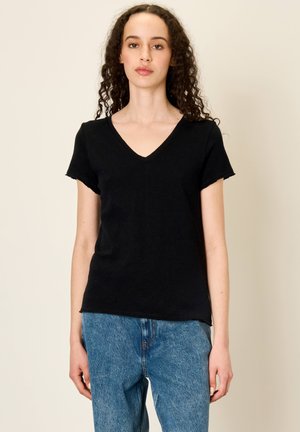 Žena kučeravými vlasmi, oblečená v čiernom krátkom tričku s výstrihom do V a modrých džínsoch, stojaca na jednoduchom svetlom pozadí.