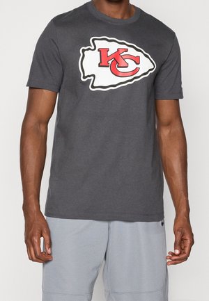 Homme portant un t-shirt gris foncé des Kansas City Chiefs avec un logo rouge et blanc et un short de sport gris clair.