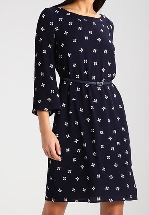 Vrouw draagt een navyblauwe jurk tot op de knie met witte vierkante patronen, driekwart mouwen en een dun bijpassende riem in de taille.