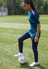 Sporty antrekk i turkis med blå mønster, marineblå bukser med rosa detaljer, og hvite fotballsko, stående på en flerfarget fotball.