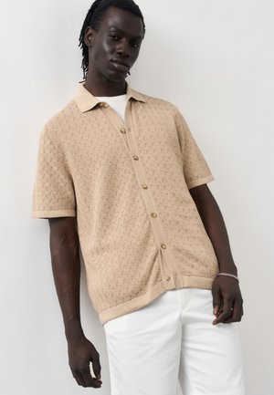 Άνδρας που φοράει μπεζ πλεκτό πουκάμισο με κοντά μανίκια και κουμπιά πάνω από λευκό μπλουζάκι και λευκό παντελόνι, στέκεται μπροστά σε απλό ανοιχτό φόντο.