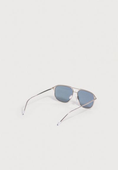 Lunettes de soleil à monture argentée avec des verres rectangulaires teintés en bleu reposant ouvertes sur une surface blanche.
