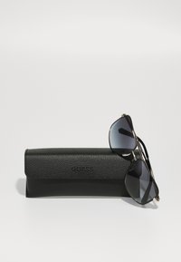 Étui à lunettes de soleil Guess texturé noir avec une paire de lunettes de soleil aviateur à monture noire reposant sur le côté droit.