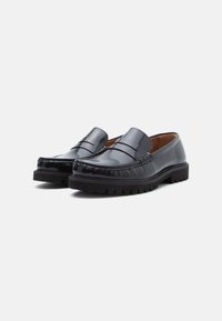 Spīdīgs melns ādas loafers pāris ar masīviem gumijas zolemi, moctoe šuvēm un viegli uzvelkamu dizainu uz vienkārša balta fona.