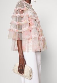 Gennemsigtig rosa bluse med flæser, blomsterprint og hvide detaljer, parret med en cremet perleclutch. Hvide bukser fuldender looket.