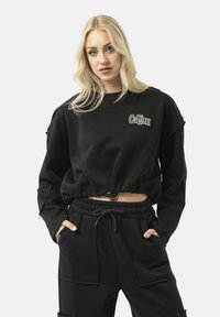 Zwarte cropped sweatshirt met een ronde hals, voorzien van een logo aan de voorkant, losse mouwen en een koord aan de onderkant. Gecombineerd met zwarte broek.