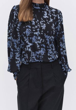 Kvinde iført en sort bluse med blå blomsterprint og lange ærmer, der er stoppet ned i højtaljede sorte bukser med hænderne i lommerne.