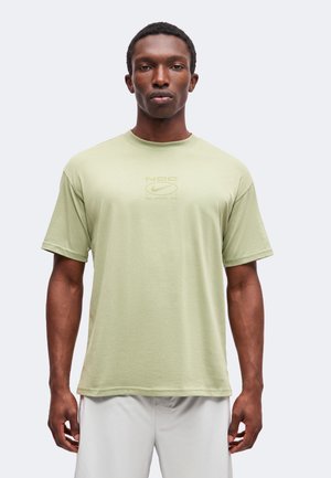 Mand står iført en lysegrøn Nike T-shirt med korte ærmer og lyse bukser mod en ensfarvet lys baggrund.
