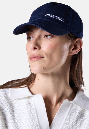 Vrouw met een marineblauwe Rossignol baseballpet en een wit textuurrijk hemd met kraag, kijkt iets naar links tegen een effen achtergrond.