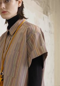 Camisa de manga curta às riscas em material suave, apresenta riscas verticais multicoloridas e um bolso no peito, combinada com um pulôver de gola alta preto por baixo.